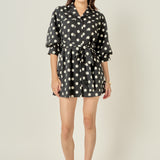 Full-length front view of a black and white polka dot shirt dress featuring 3/4 blouson sleeves, a notched collar, and a cinched tie belt sash.