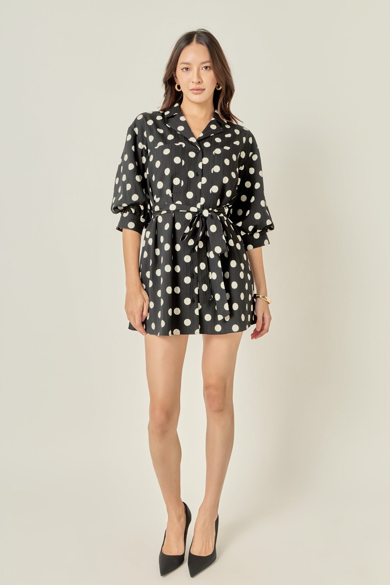 Full-length front view of a black and white polka dot shirt dress featuring 3/4 blouson sleeves, a notched collar, and a cinched tie belt sash.