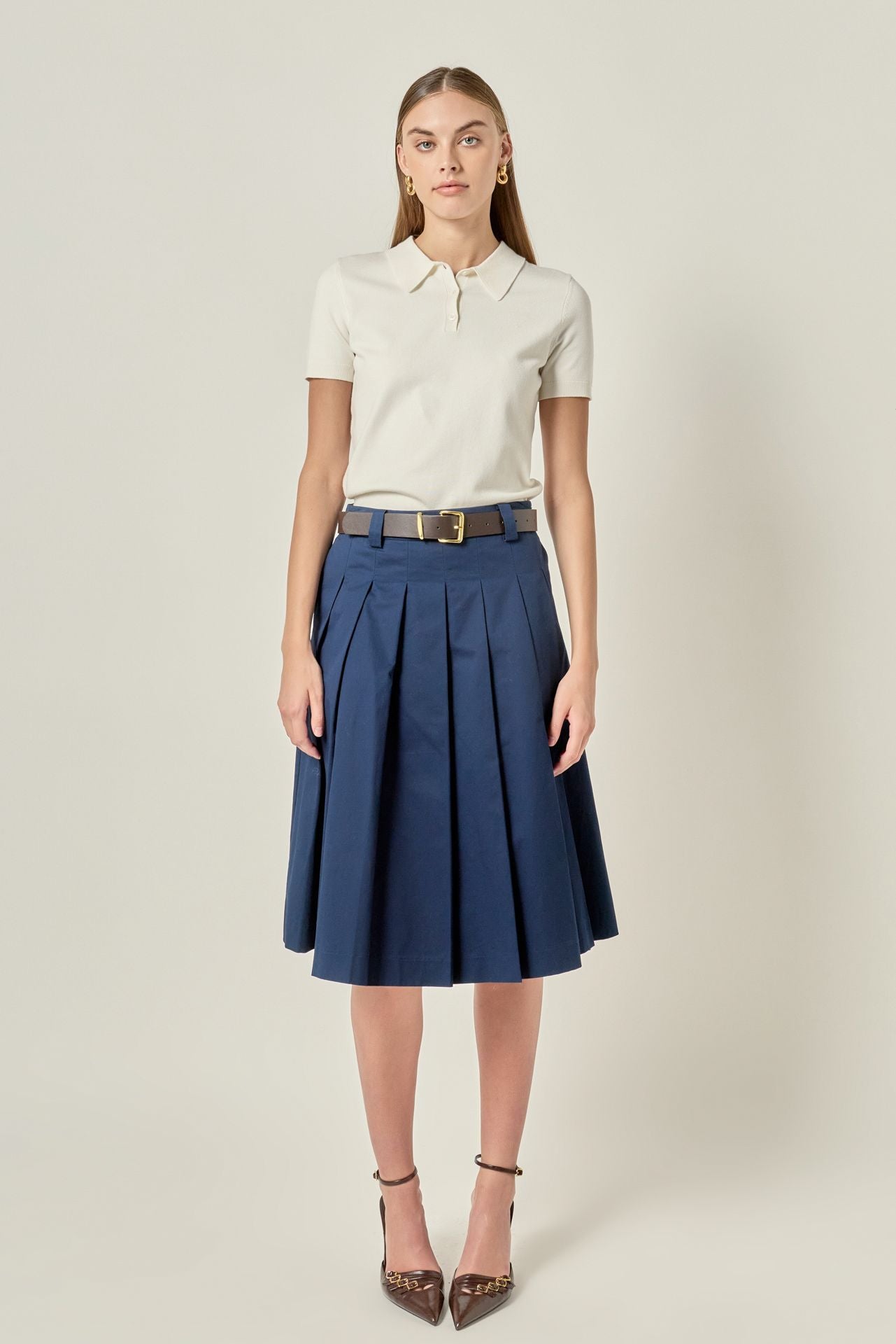 Full-length front view of a navy blue pleated cotton midi skirt featuring belt loops, styled with a cream short-sleeve polo shirt.