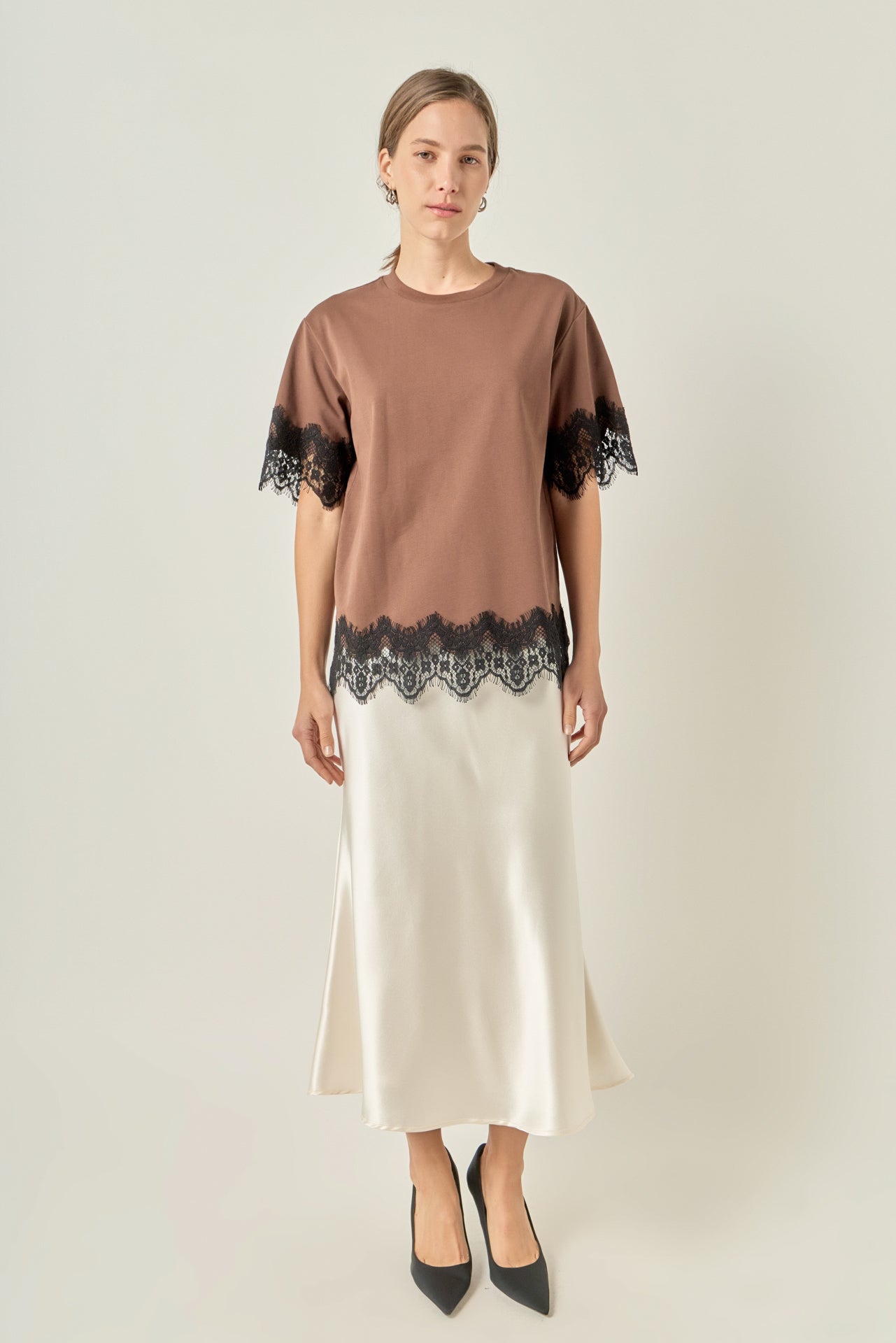 Full-length front view of a brown short-sleeve top featuring black contrast eyelash lace trim at the sleeves and hem, styled with a cream satin midi skirt.