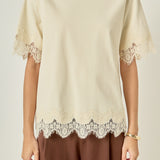 Upper body close-up view of a cream round-neck top highlighting the delicate cream lace edging along the sleeves and hem.