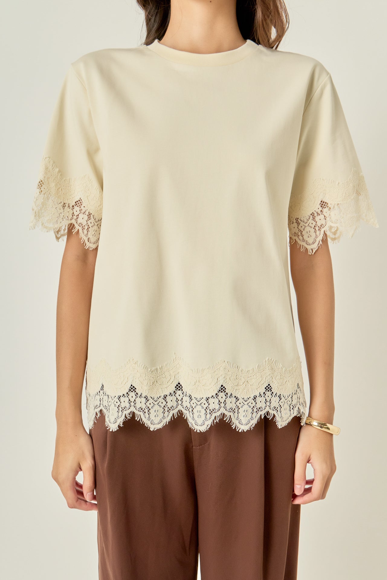 Upper body close-up view of a cream round-neck top highlighting the delicate cream lace edging along the sleeves and hem.
