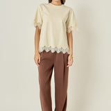 Full-length front view of a cream short-sleeve top featuring matching tonal cream eyelash lace trim at the cuffs and hem, styled with brown tailored trousers.