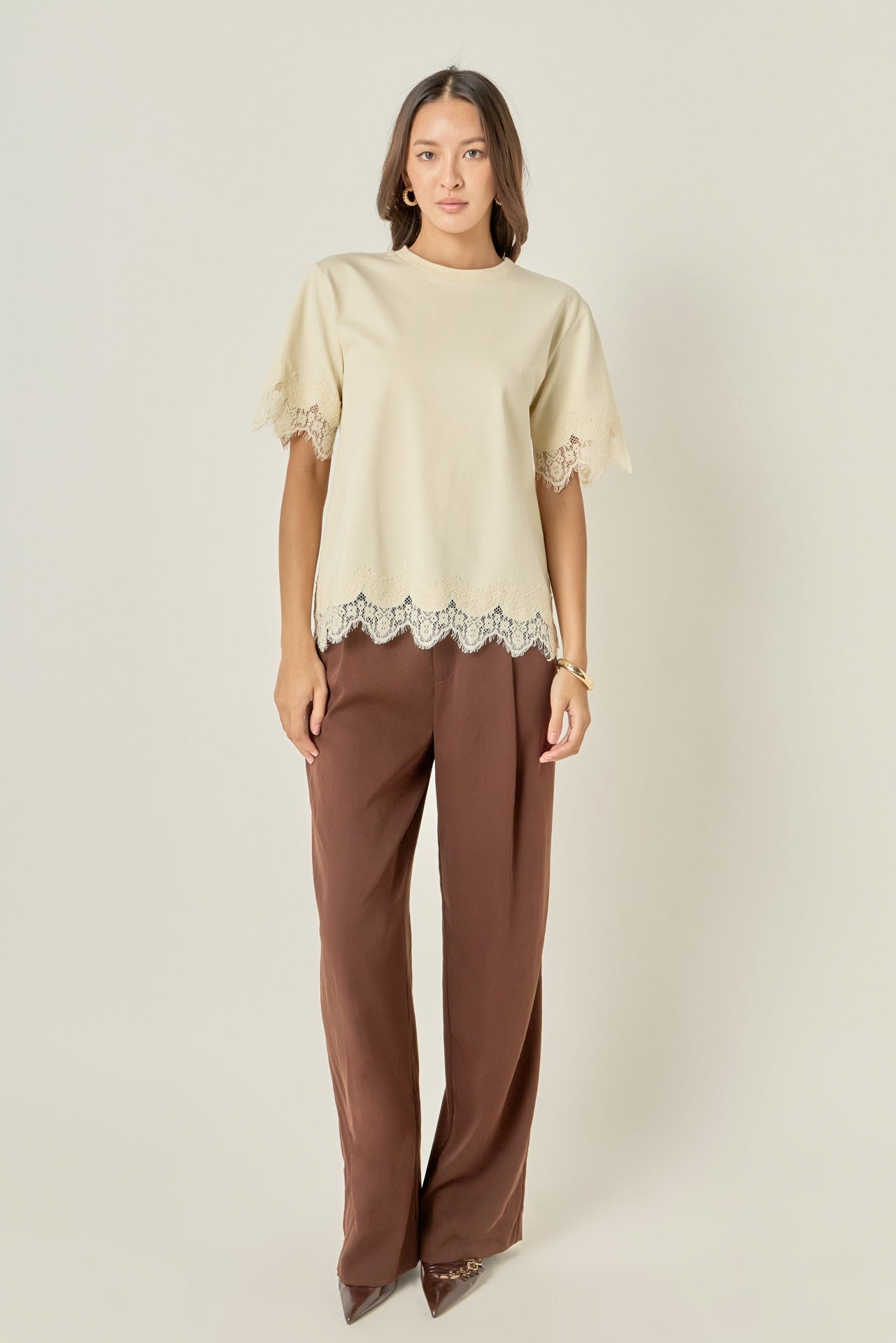 Full-length front view of a cream short-sleeve top featuring matching tonal cream eyelash lace trim at the cuffs and hem, styled with brown tailored trousers.