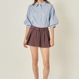 Full-length front view of a dusty blue striped button-down shirt featuring voluminous balloon sleeves, styled tucked into a brown pleated mini skirt.