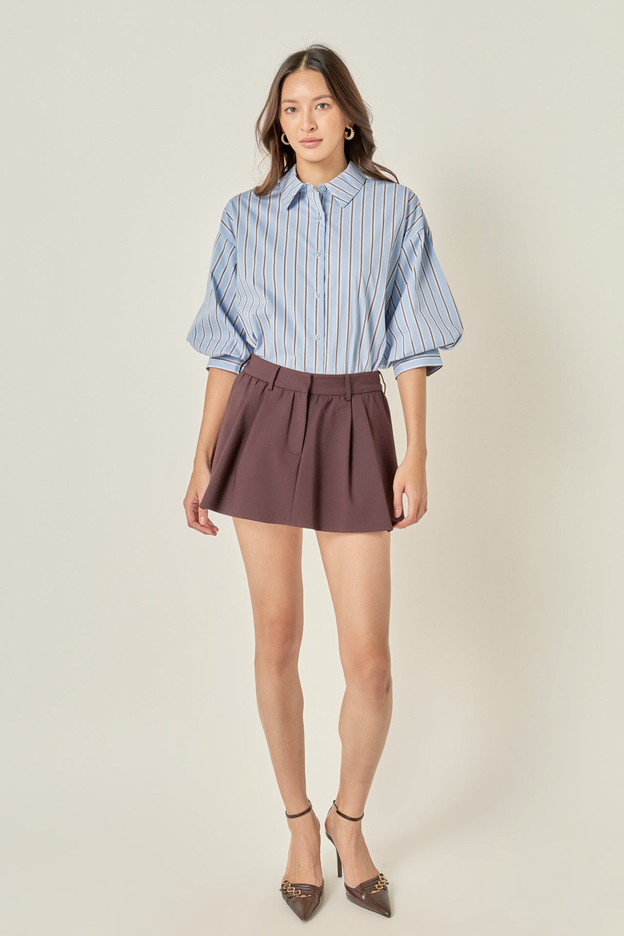 Full-length front view of a dusty blue striped button-down shirt featuring voluminous balloon sleeves, styled tucked into a brown pleated mini skirt.